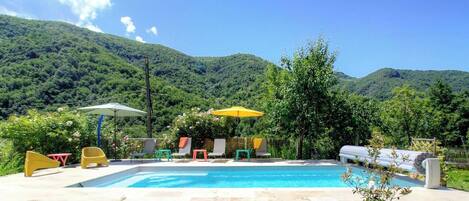 Une piscine entourée d’une végétation luxuriante et de montagnes, avec des chaises longues et des parasols.