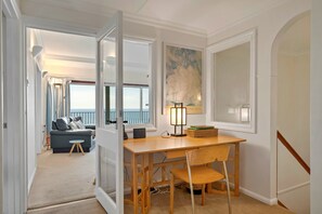 Ein Zimmer mit einem Holztisch, einem Stuhl, einer Lampe, einem Gemälde, einem Sofa und Meerblick durch ein Fenster.