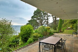 Um pátio com mesa e cadeiras, rodeado por vegetação e vista para o mar.