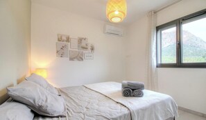 A bedroom with a bed, pillows, a rolled towel, wall art, a hanging light fixture, an air conditioning unit, and a window with a view. - Villa l'alivu - AFA - Gîte (AFA)