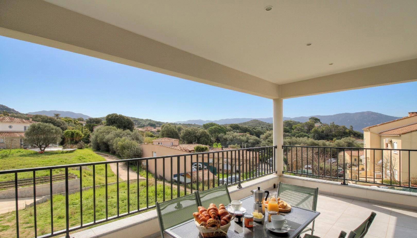 Un balcon avec une table préparée pour le petit-déjeuner, d’où l’on peut admirer un paysage verdoyant et des collines lointaines.