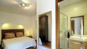 Un dormitorio con cama, lámparas de noche, cabecero de madera y una puerta que da a un baño con ducha, lavamanos y espejo.