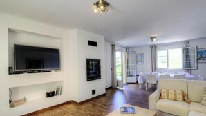 Una sala de estar con una televisión de pantalla plana, una estantería, un sofá y una zona de comedor con una mesa y sillas.