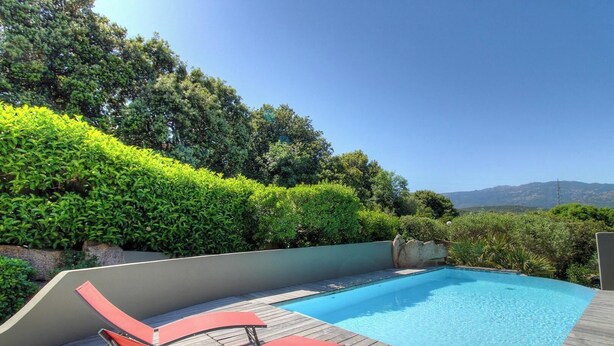 Piscina com deck de madeira, cadeiras de descanso vermelhas e vista para árvores e montanhas.