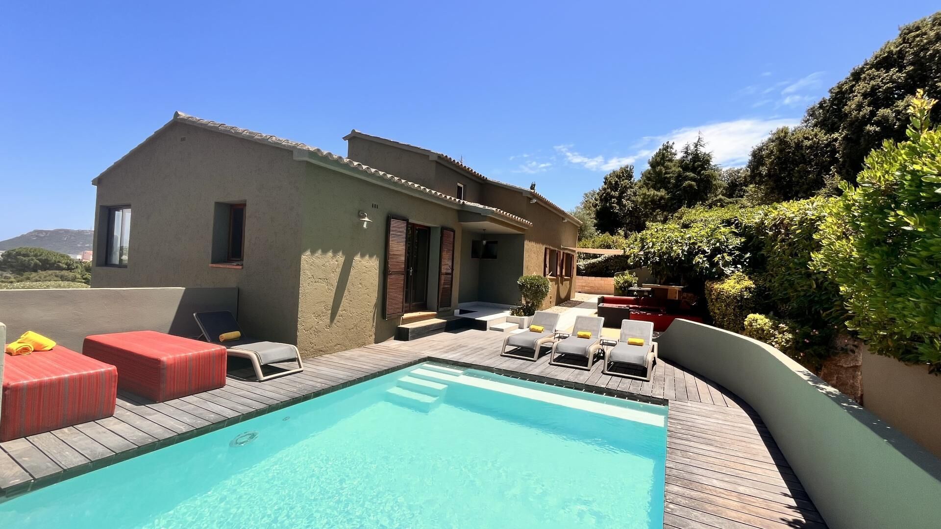 Un espace aménagé au bord de la piscine, avec des chaises longues, une terrasse en bois et un bâtiment doté d’un toit recouvert de tuiles.