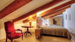 Quarto com teto de madeira, uma cama, uma cadeira vermelha, uma mesinha e um criado-mudo.