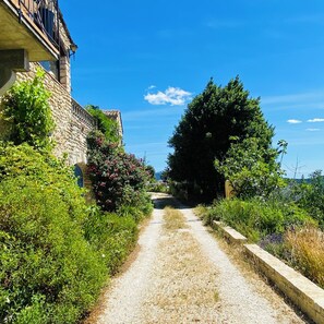 Ein Kiesweg führt zu einem Steinhaus mit Balkon, umgeben von üppiger Vegetation und blauem Himmel.