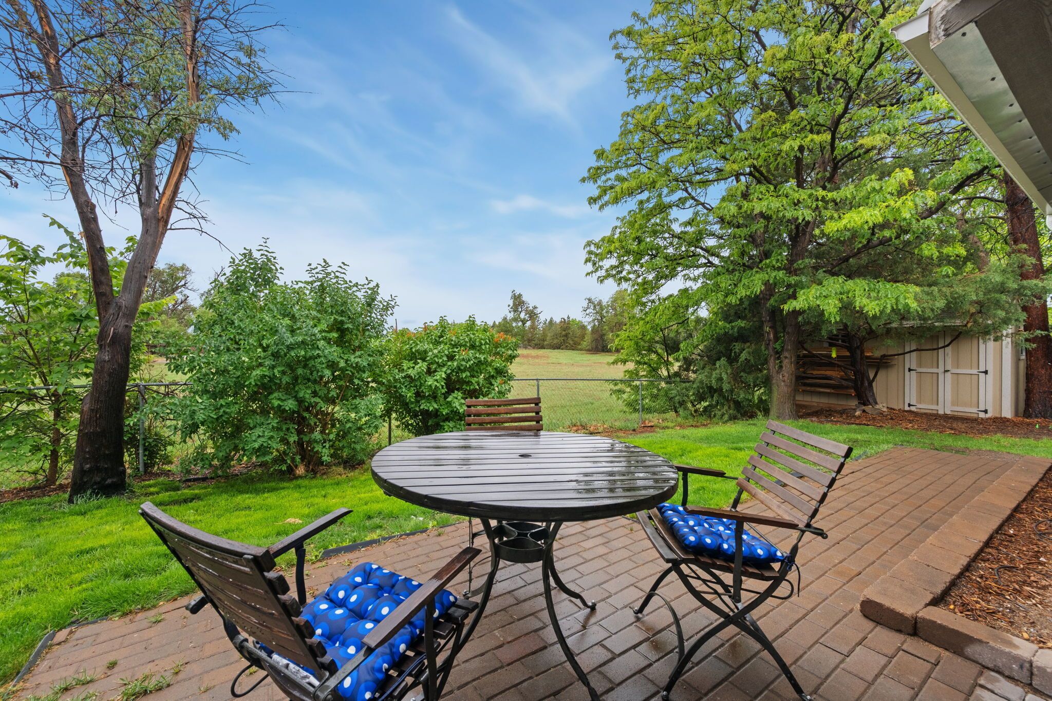An outdoor patio with a round table and two chairs, surrounded by greenery and trees.