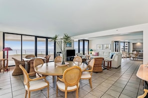 Ein großzügiger Wohnbereich mit Esstisch, Korbstühlen, einer Couch und einem Fernseher. Große Fenster bieten Blick auf das Meer.