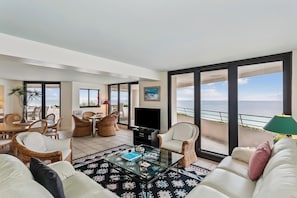 Ein modernes Wohnzimmer mit einem Glastisch, weißen Sofas, Korbstühlen und einem großen Fernseher.
