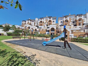 Ein Spielplatz mit Schaukeln und Rutsche, umgeben von einem gepflegten Rasen und einem Gebäude mit mehreren Balkonen.