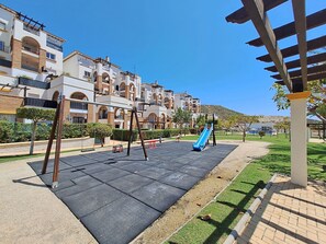 Ein Spielplatz mit Schaukeln und Rutsche, umgeben von einem gepflegten Rasen und einem Gebäude mit Balkonen.