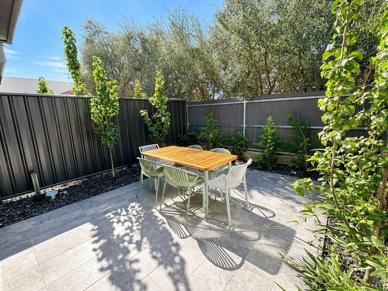 An outdoor patio with a wooden table and metal chairs, surrounded by a black fence and greenery.
