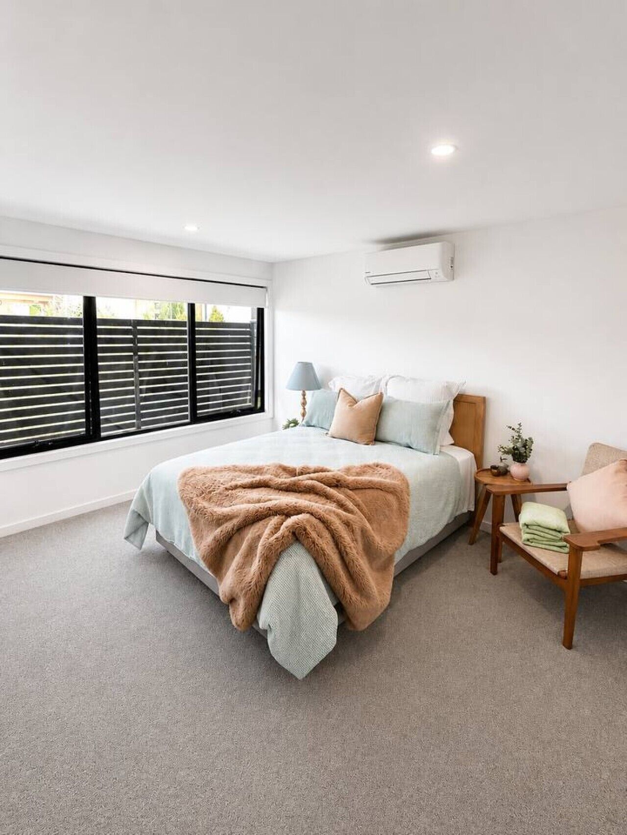 A bedroom with a large window, a bed with a blanket, a bedside table with a lamp, and a small plant.