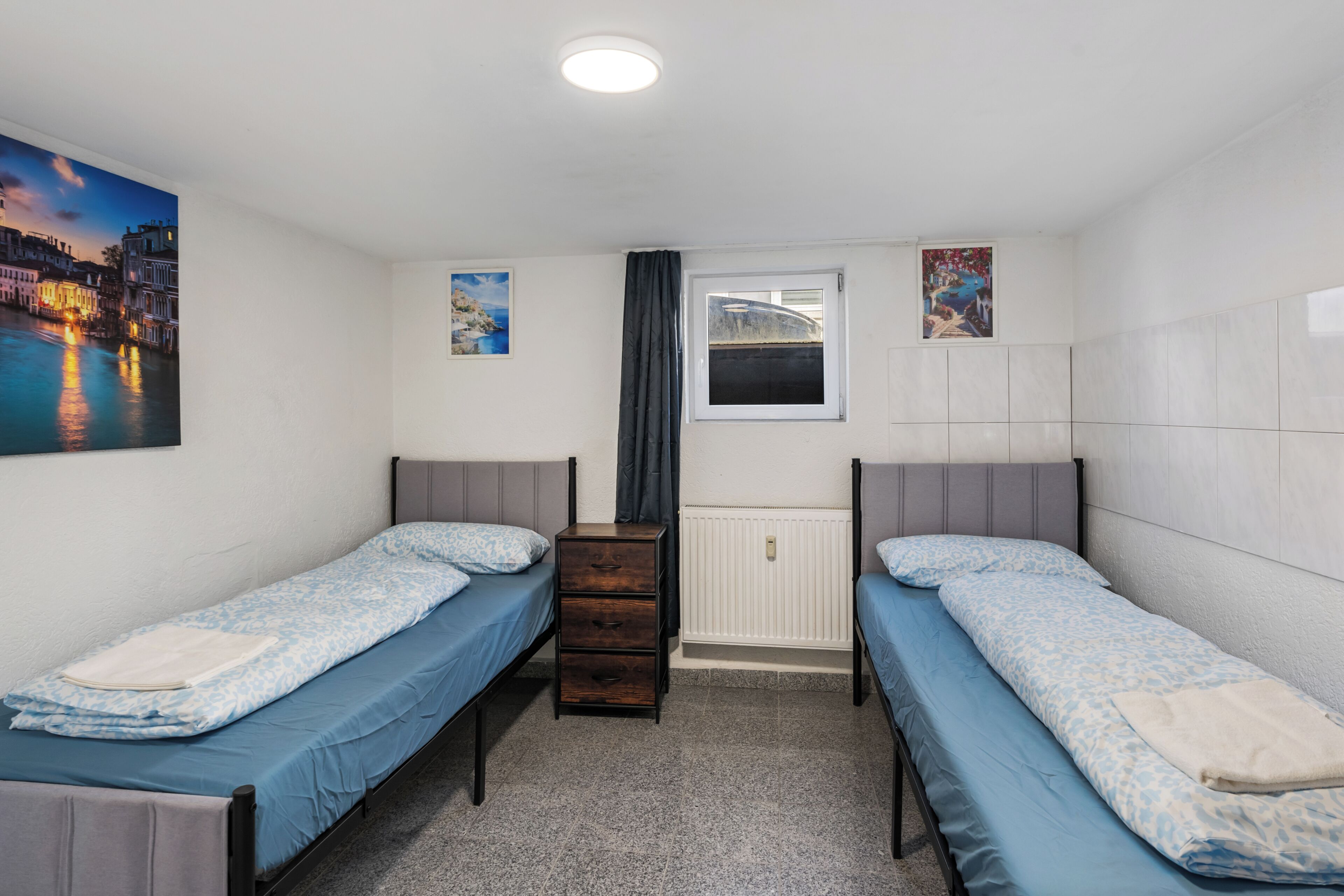 A room with two single beds, a dresser, a window with curtains, and a radiator.