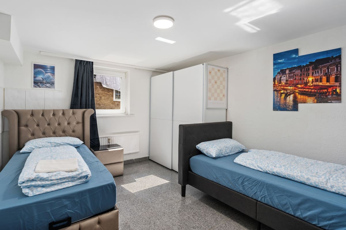 Una habitación con dos camas, una ventana con cortinas, un radiador, una obra de arte colgada en la pared y un estampado de cuadros en la pared.