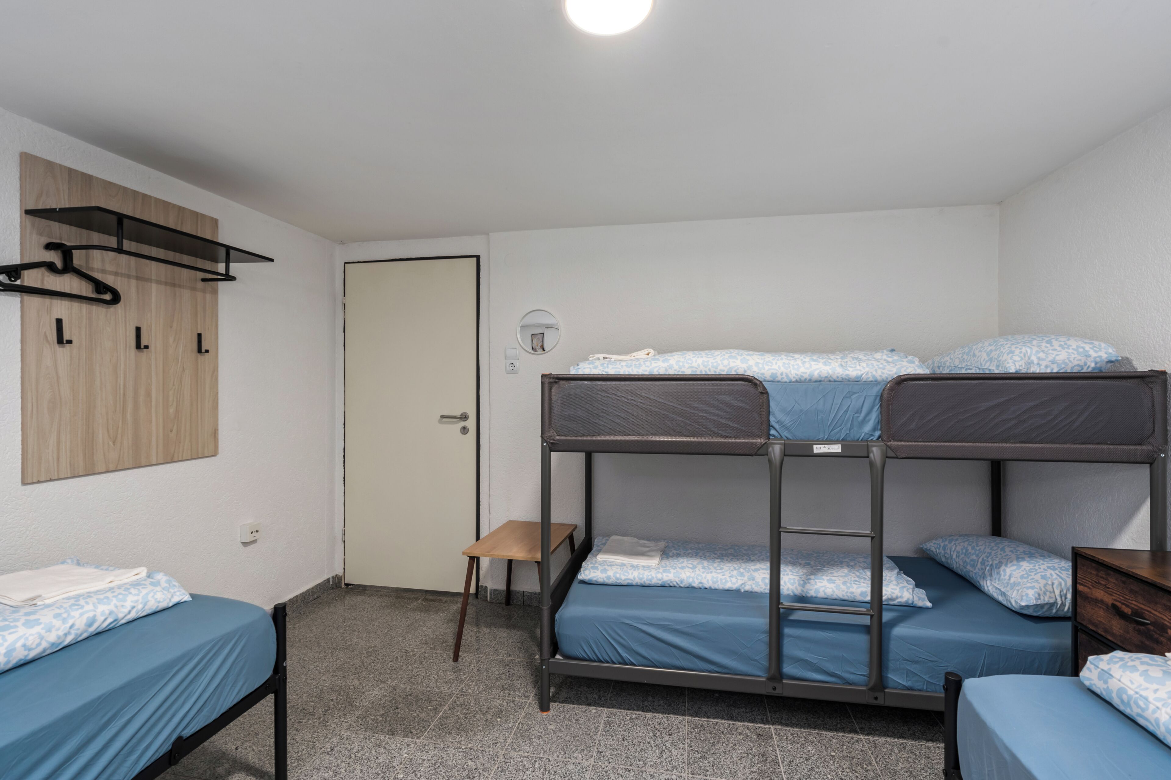 Un dormitorio con literas, un estante de madera con perchas, un taburete pequeño de madera y una puerta.