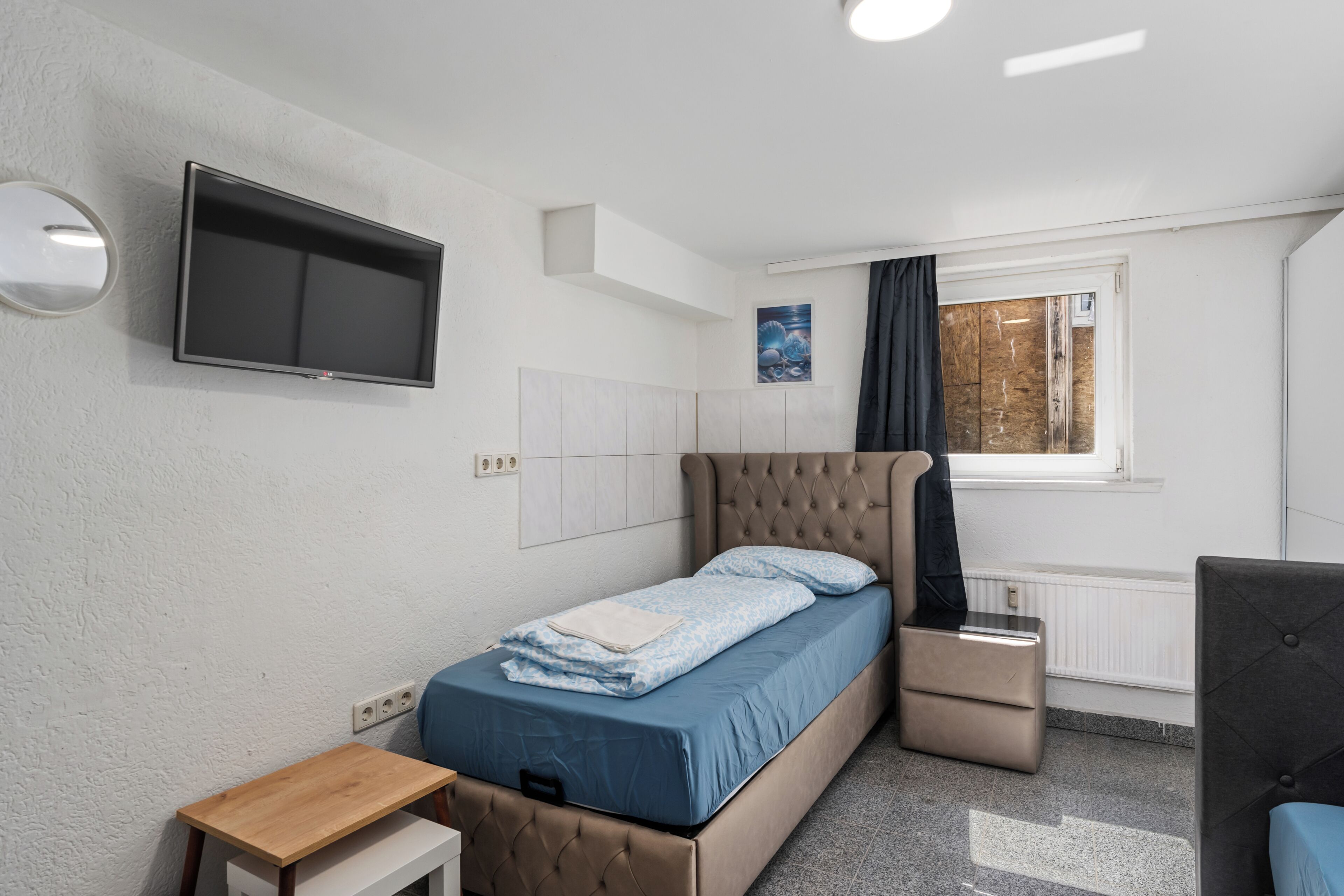 Un dormitorio con cama, televisor, una mesita, ventana con cortinas y radiador.