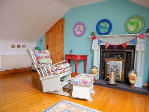A room with a wooden floor, a white rocking chair, a small table, and a fireplace with decorative items. - House of the Sea - Camp (Kilgobban)