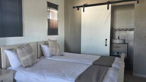 Cama doble con dos almohadas, mesita de noche, ventana con persianas, toallero y baño con lavabo y espejo.