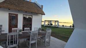 Un comedor con una mesa de madera y sillas blancas, un techo de paja y vistas a una masa de agua tranquila.
