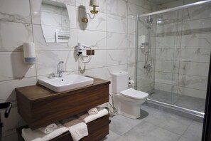Un baño con pared de azulejos de mármol, lavamanos blanco, inodoro, ducha con mampara de vidrio y tocador de madera con toallas.