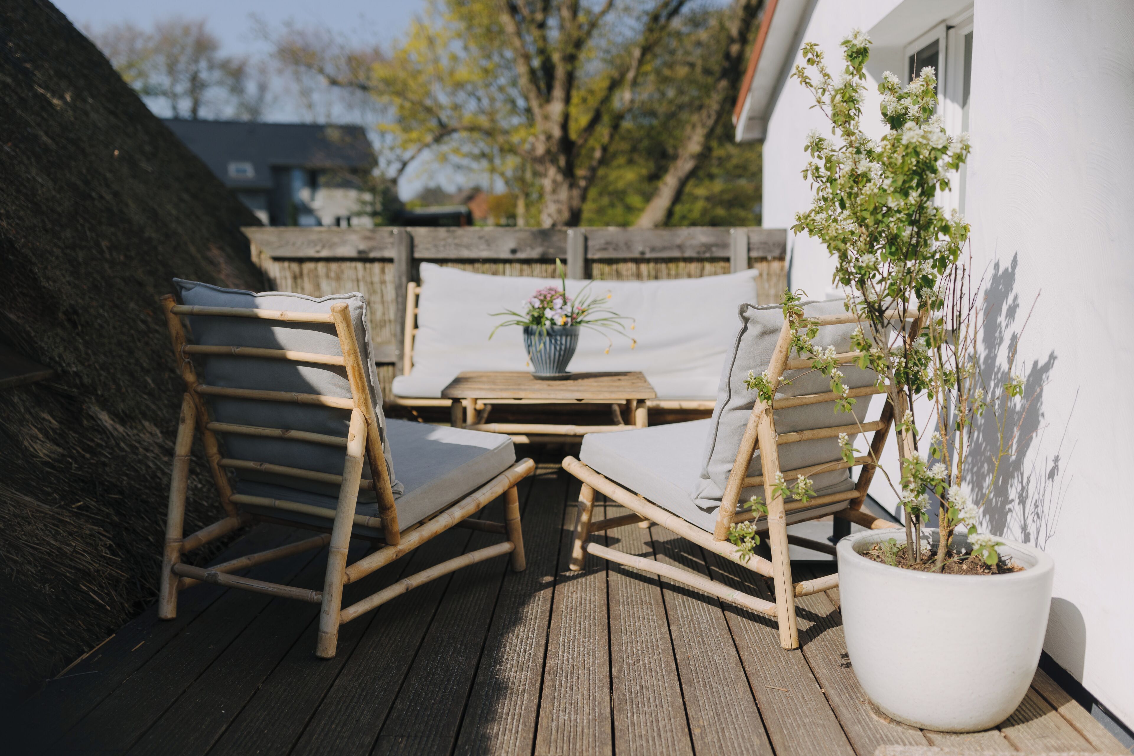 Een houten terras met twee rieten stoelen en een tafel, een bloempot en een witte muur.