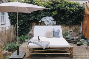 Ein Bett mit Kissen, einer Decke und einer Weinflasche auf einer Holzplattform, unter einem großen Sonnenschirm, umgeben von Grünpflanzen und einer Steinmauer.