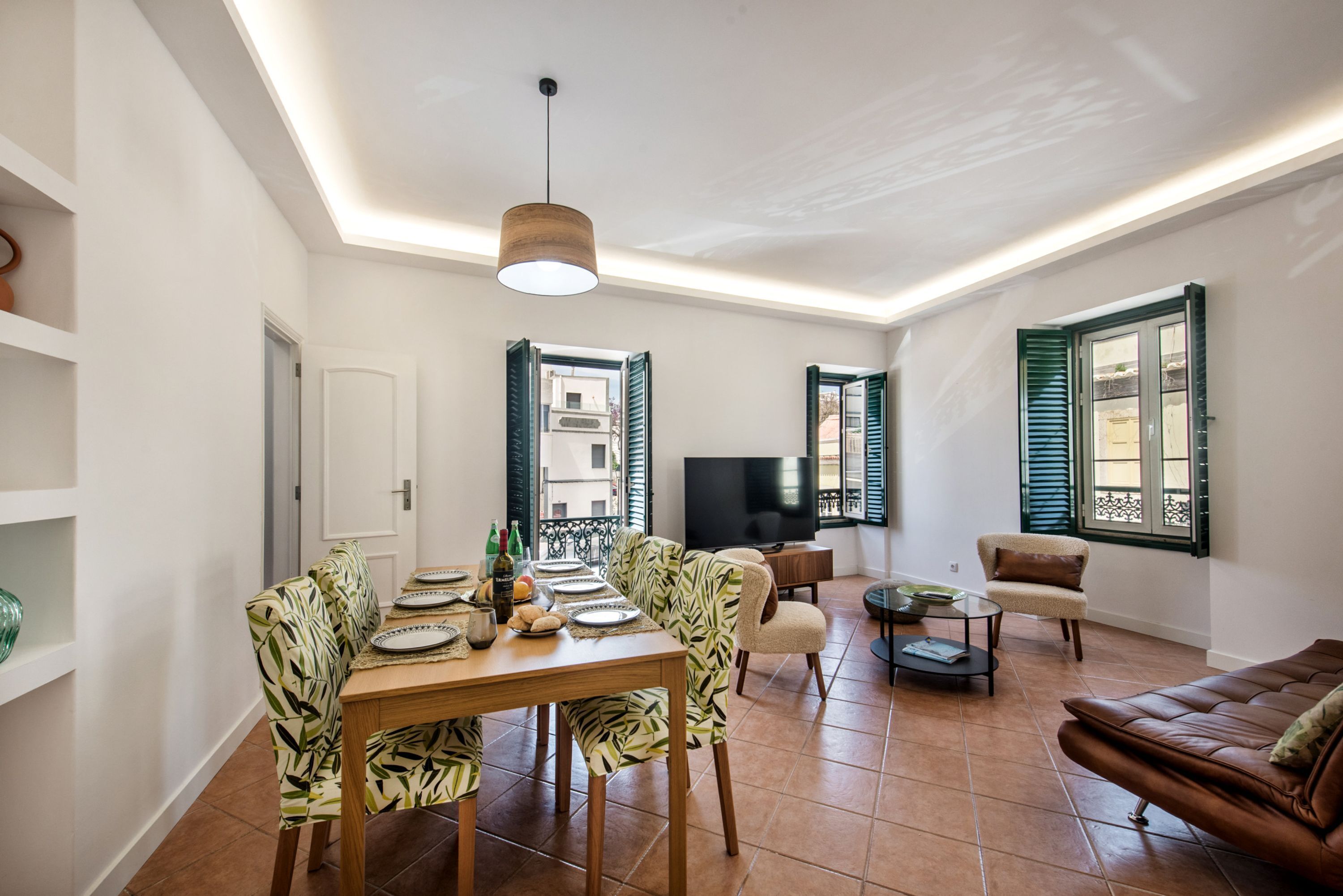 A dining area with a table set for four, a sofa, a TV, and a balcony with shutters.