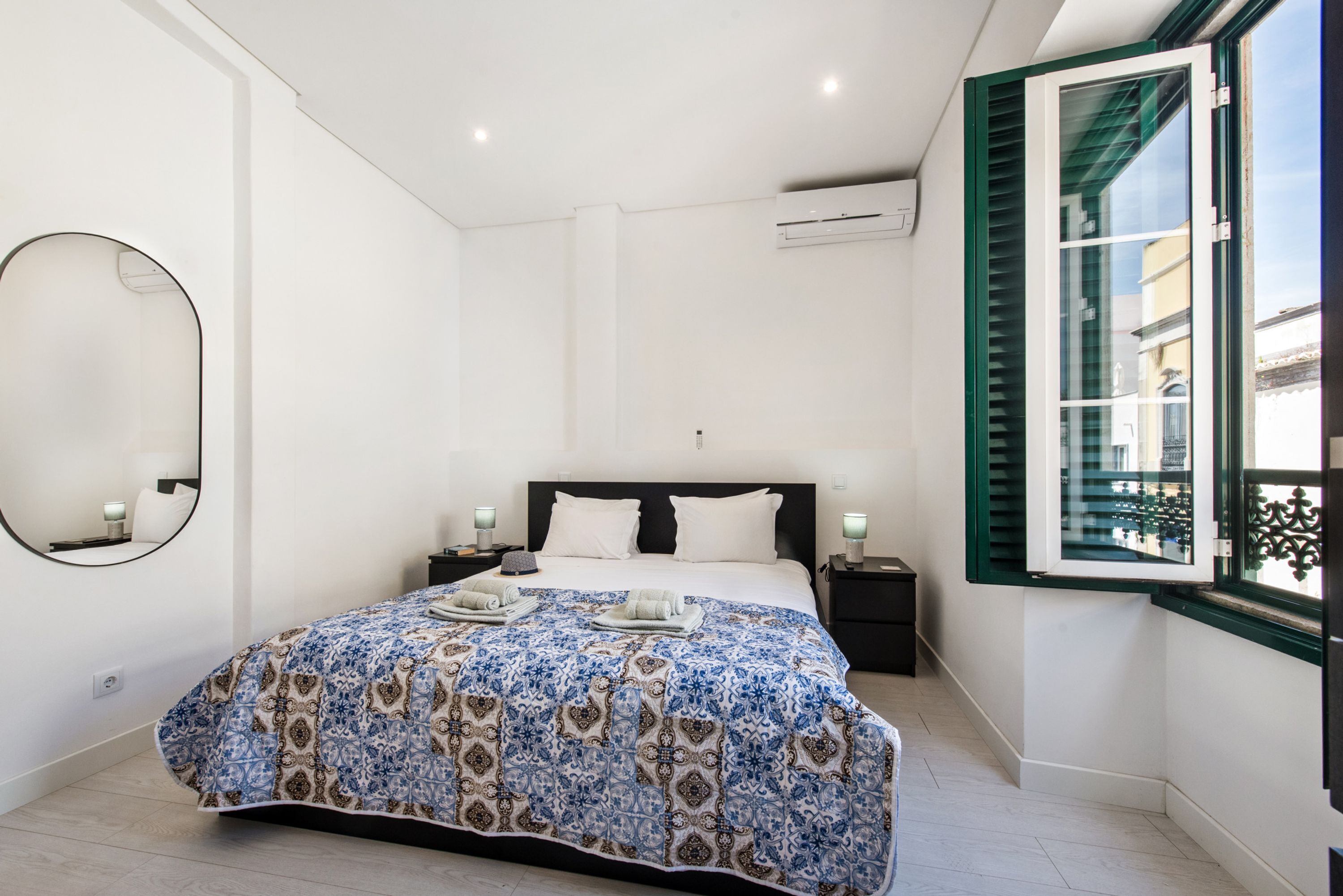 Quarto com cama, mesas de cabeceira, espelho e janela com persianas.