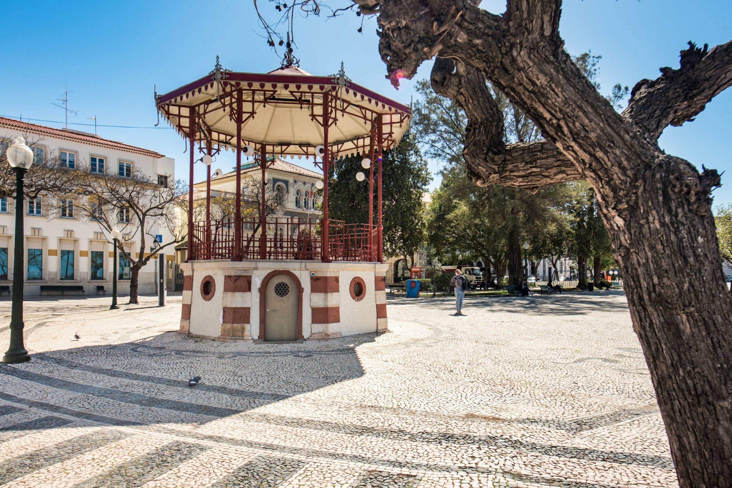 A gazebo with a tiled floor, a building with a tiled roof, and a tree in the foreground.