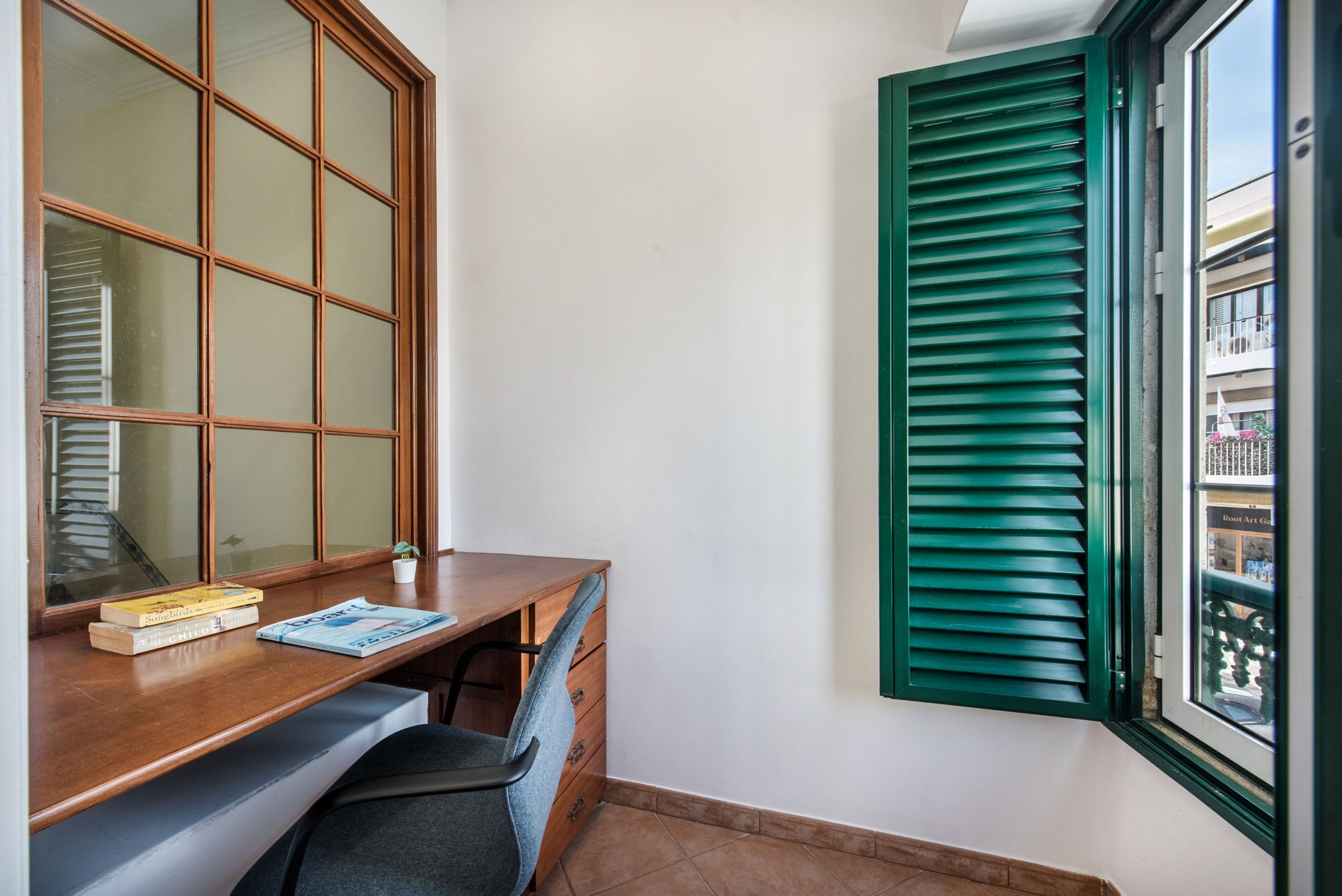 A corner room with a wooden desk, a chair, a window with green shutters, and a view of buildings outside.
