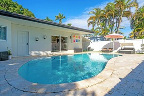Ein Pool mit geschwungener Kante, ein weißes Gebäude mit Tür und Fenstern sowie Palmen im Hintergrund.