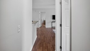 A hallway with white walls and wooden flooring leading to a kitchen area with a TV mounted on the wall.