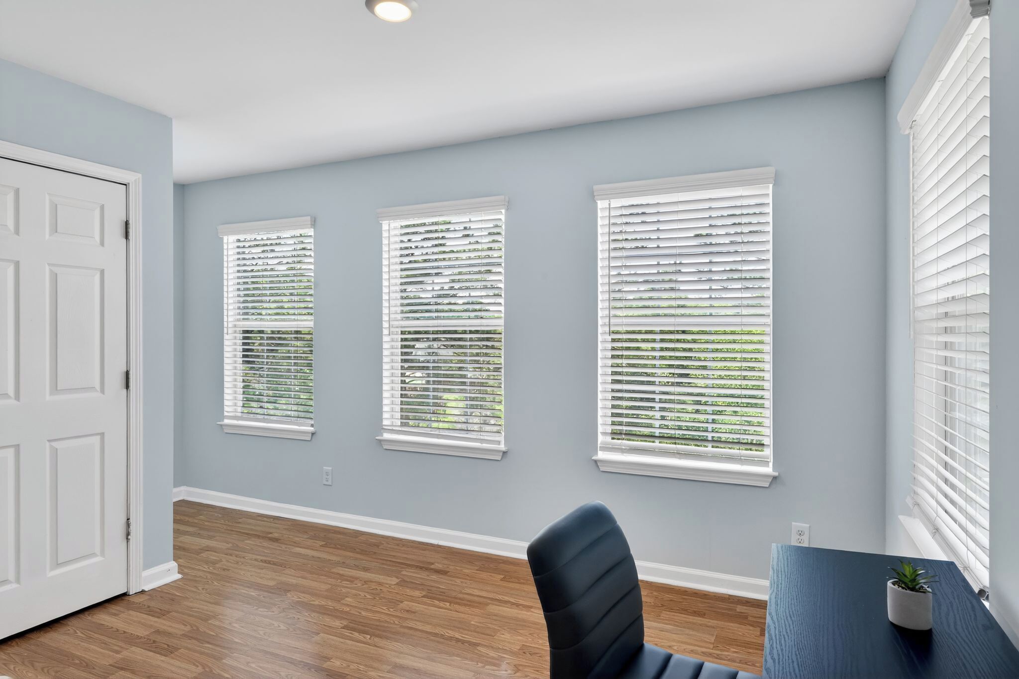A room with three windows, a door, a table with a plant, and a chair.