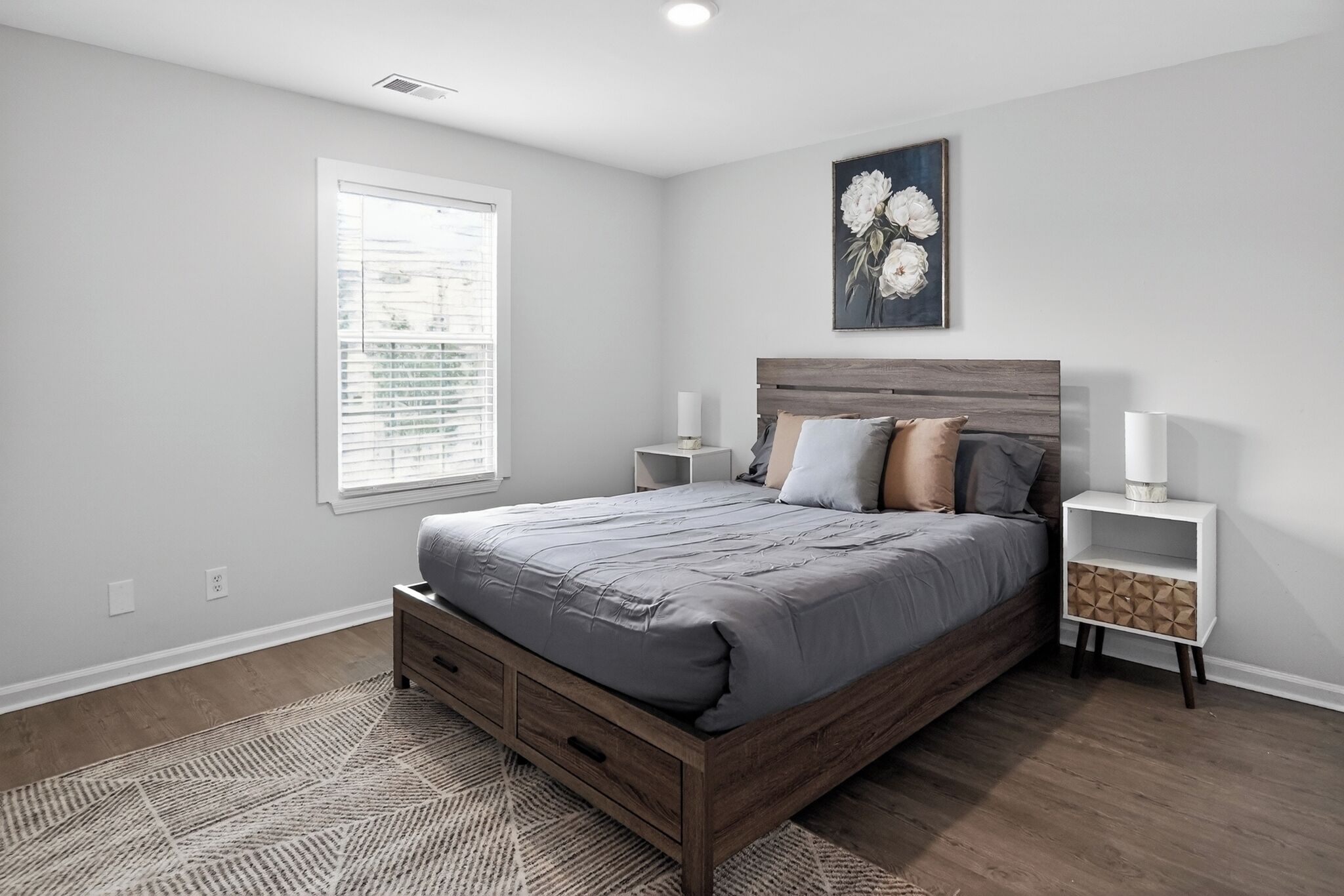 Une chambre à coucher avec un grand lit, des tables de chevet, une fenêtre avec des stores et un tableau accroché au mur.