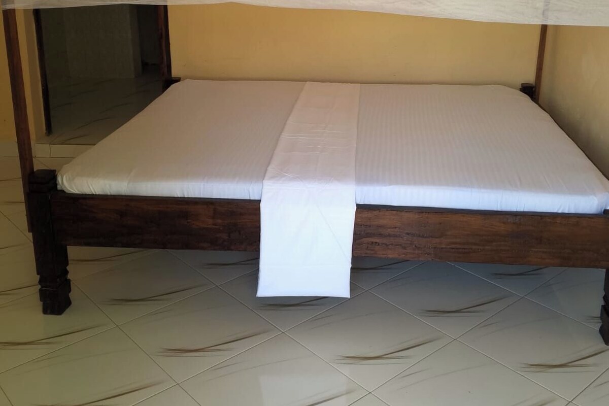 Una cama con estructura de madera y ropa de cama blanca, una mosquitera colgando sobre ella, y un piso de baldosas.