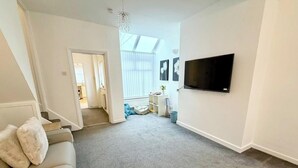 Una sala de estar con alfombra gris, sofá blanco, televisor montado en la pared y una escalera que lleva a otra habitación.