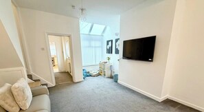 Ein Wohnzimmer mit grauem Teppich, einem weißen Sofa, einem an der Wand befestigten Fernseher und einer Treppe, die in einen anderen Raum führt.