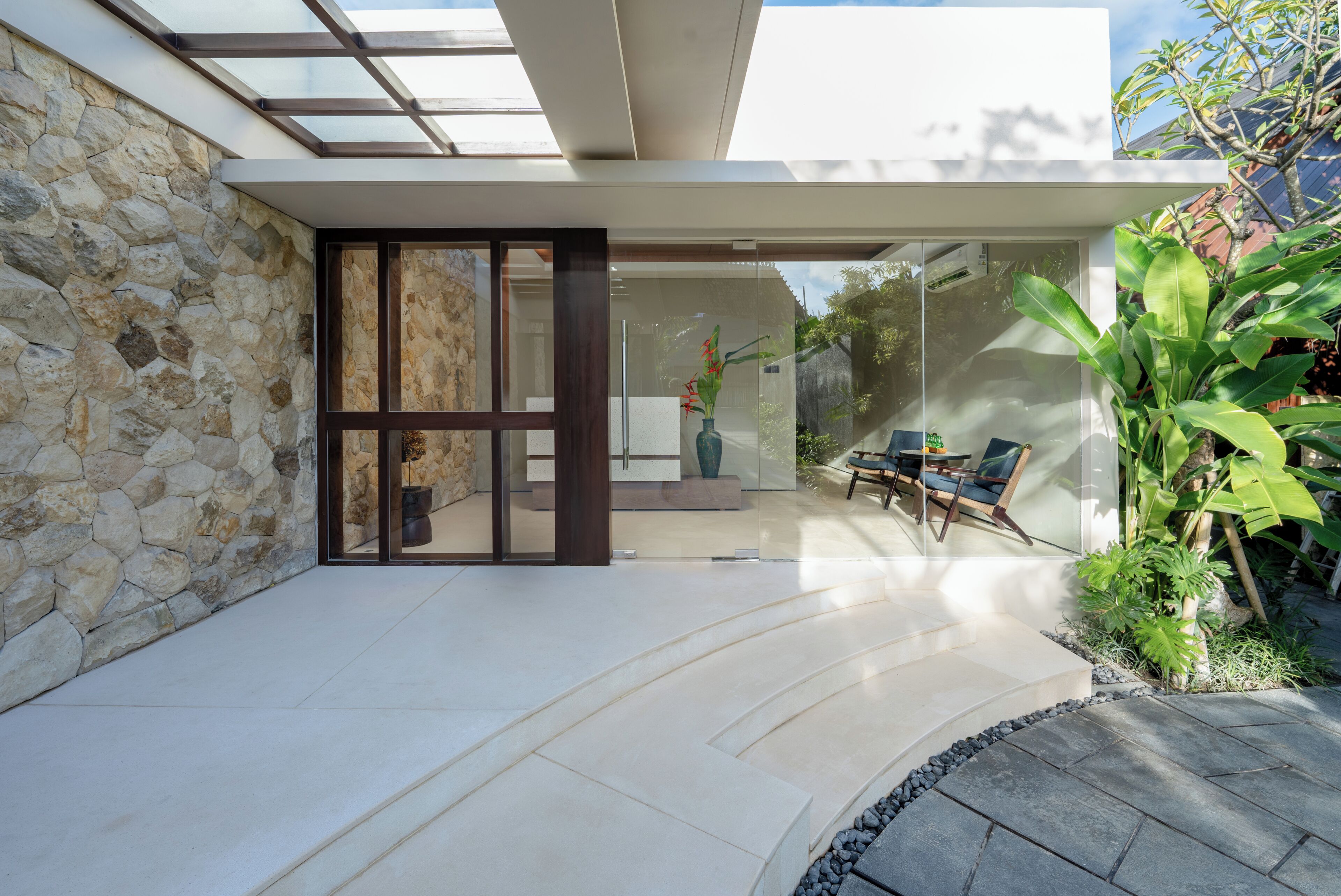 A modern house with a stone exterior, a glass door leading to a patio with outdoor furniture, and a garden with tropical plants.