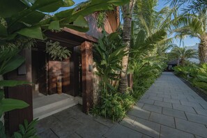 Un camino que lleva a un edificio de madera con una puerta, rodeado de plantas tropicales y palmeras.