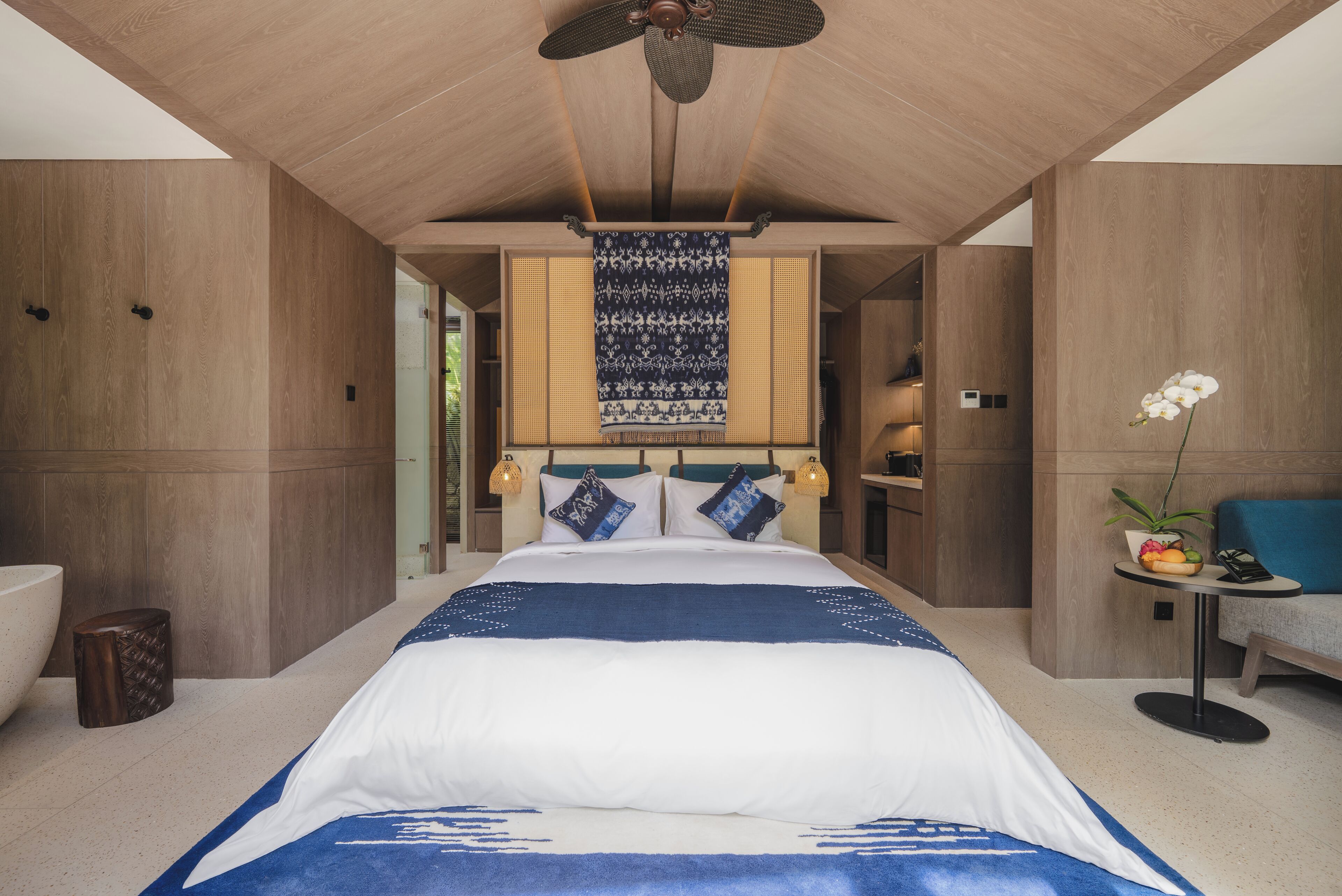 A modern hotel room with a large bed, a ceiling fan, a wall-mounted tapestry, and a small table with a plant.