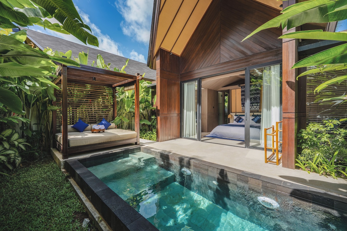 Zona junto a la piscina con terraza de madera, área de estar con cojines y una sala con paredes de vidrio.