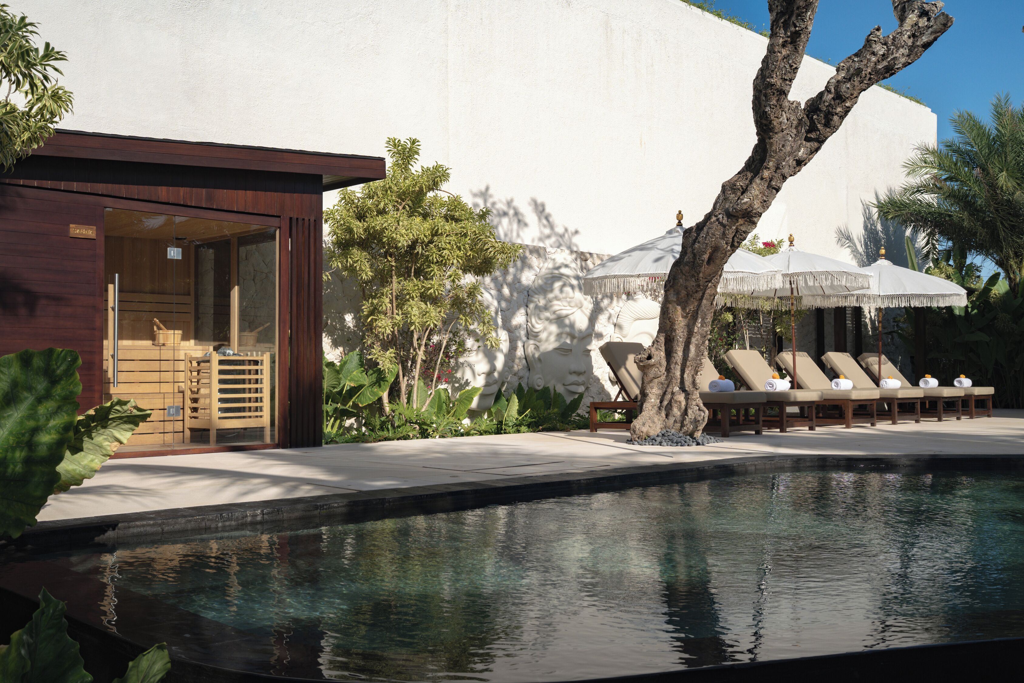 A poolside area with lounge chairs, a wooden structure with a glass door, and a white wall with a face sculpture.