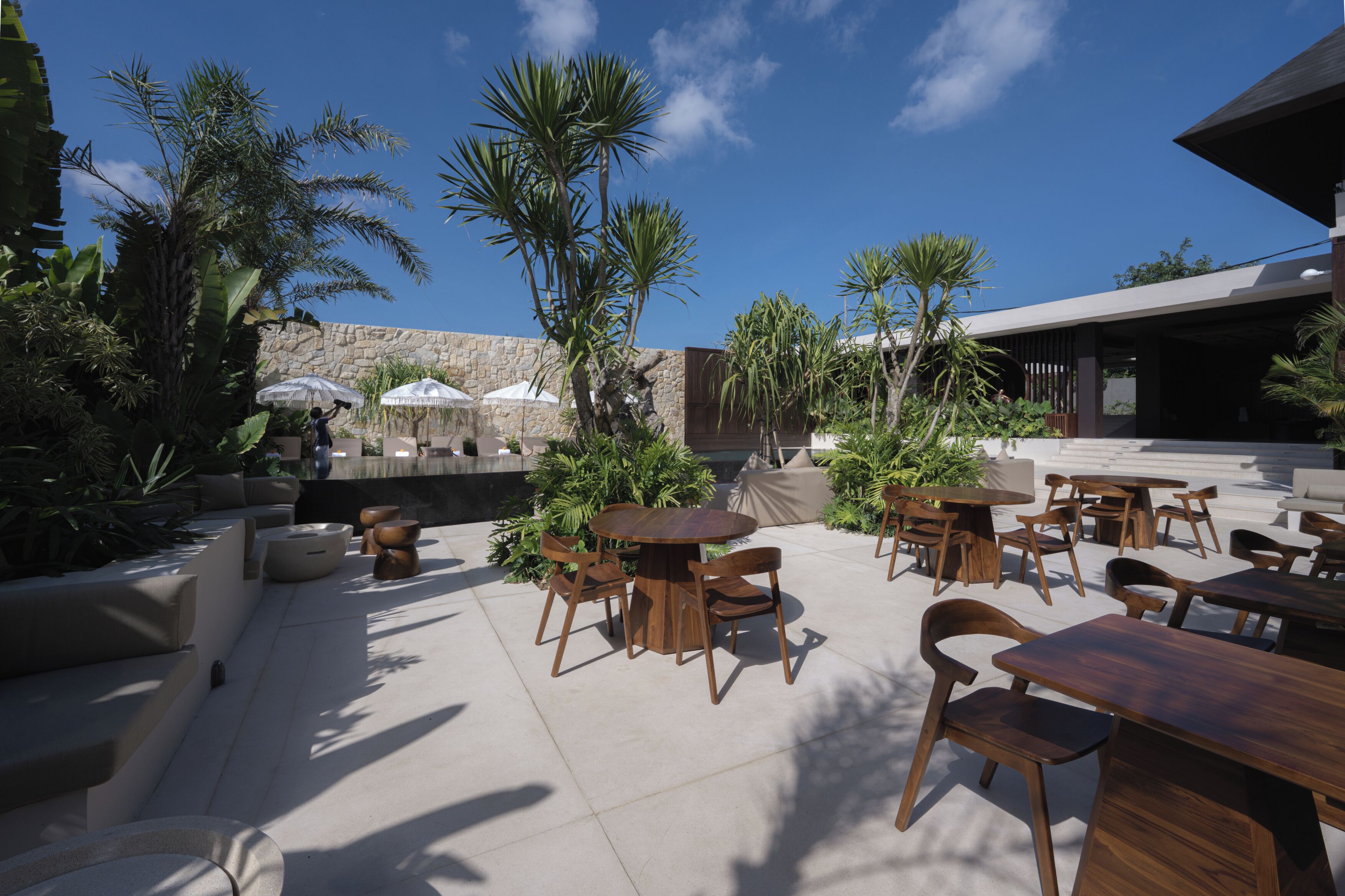 An outdoor patio area with multiple wooden tables and chairs, surrounded by various plants and umbrellas.