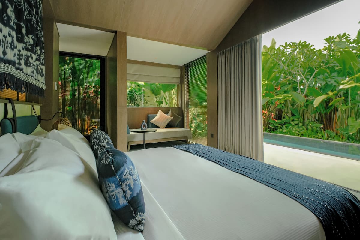 A modern bedroom with a large bed, a seating area, and a view of greenery outside.