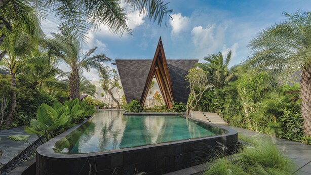 Una piscina rodeada de abundante vegetación y un edificio moderno con techo triangular.