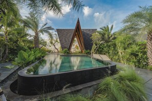 Una piscina envoltada de vegetació exuberant i un edifici modern amb un teulat triangular.