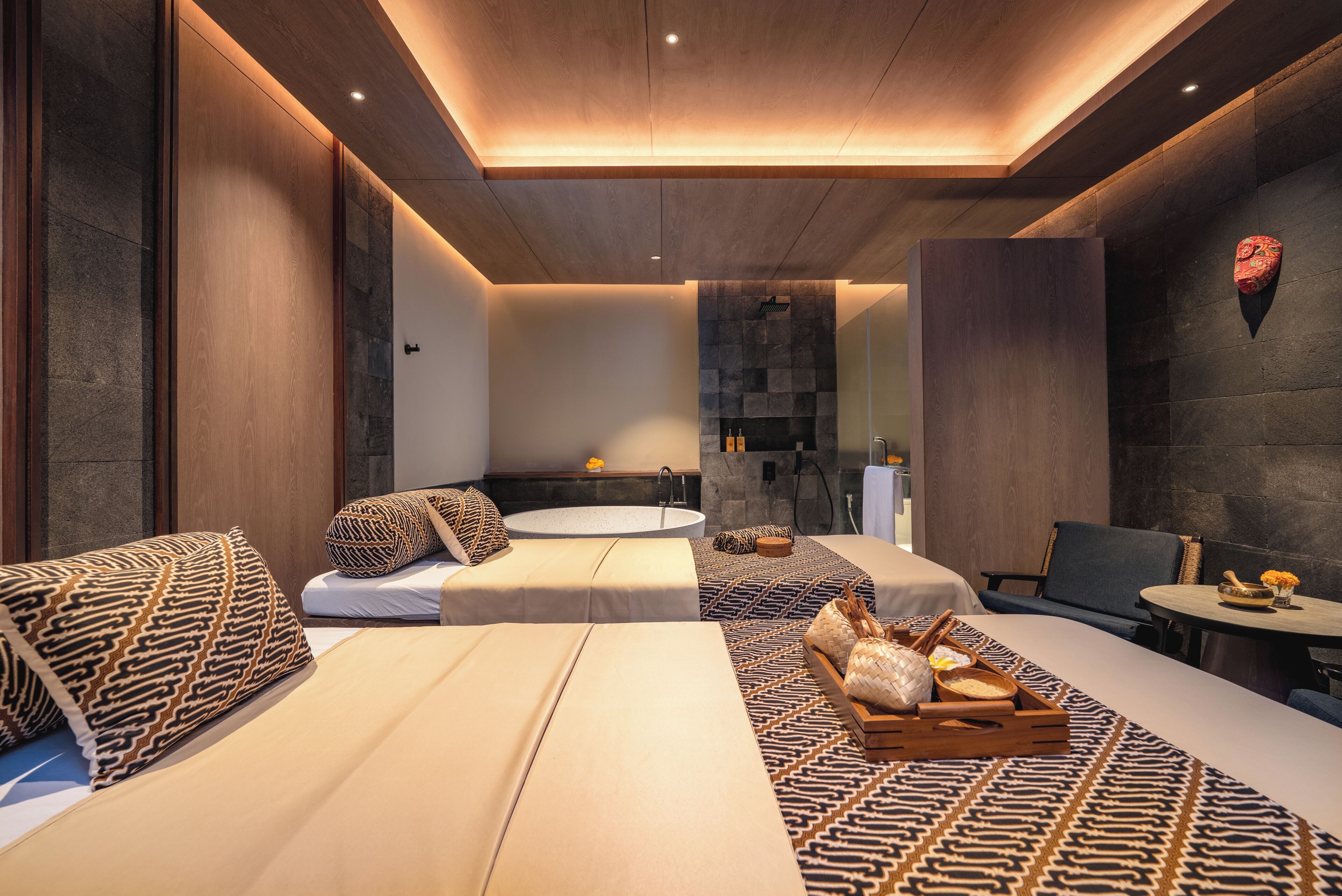 A modern spa room with a large bathtub, two seating areas with patterned cushions, and a small table with a bowl and flowers.