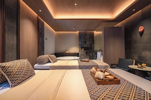 A modern spa room with a large bathtub, two seating areas with patterned cushions, and a small table with a bowl and flowers. - Nuju Uluwatu - Adults Only (Pecatu)